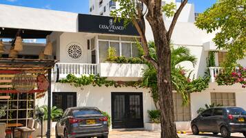 Um hotel com fachada em preto e branco, mesas ao ar livre e uma árvore em primeiro plano.