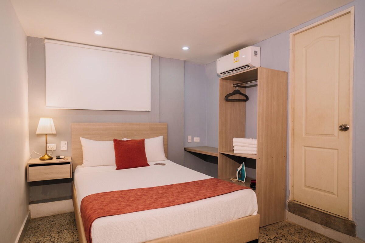 Una habitación de hotel con cama, mesita de noche con lámpara, armario y puerta.