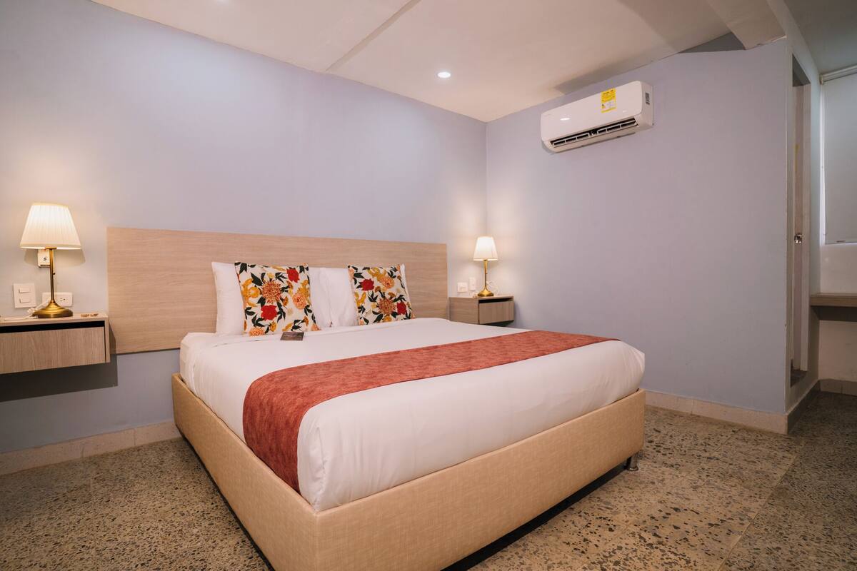 Een hotelkamer met een groot bed, twee nachtkastjes met lampen, een airconditioning en een deur aan de rechterkant.