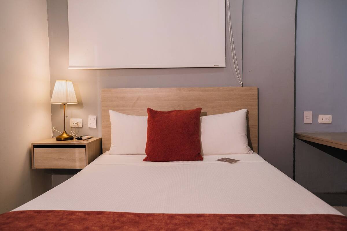 Een net opgemaakt bed met witte lakens en een rood kussen, een bedlampje en een houten nachtkastje.