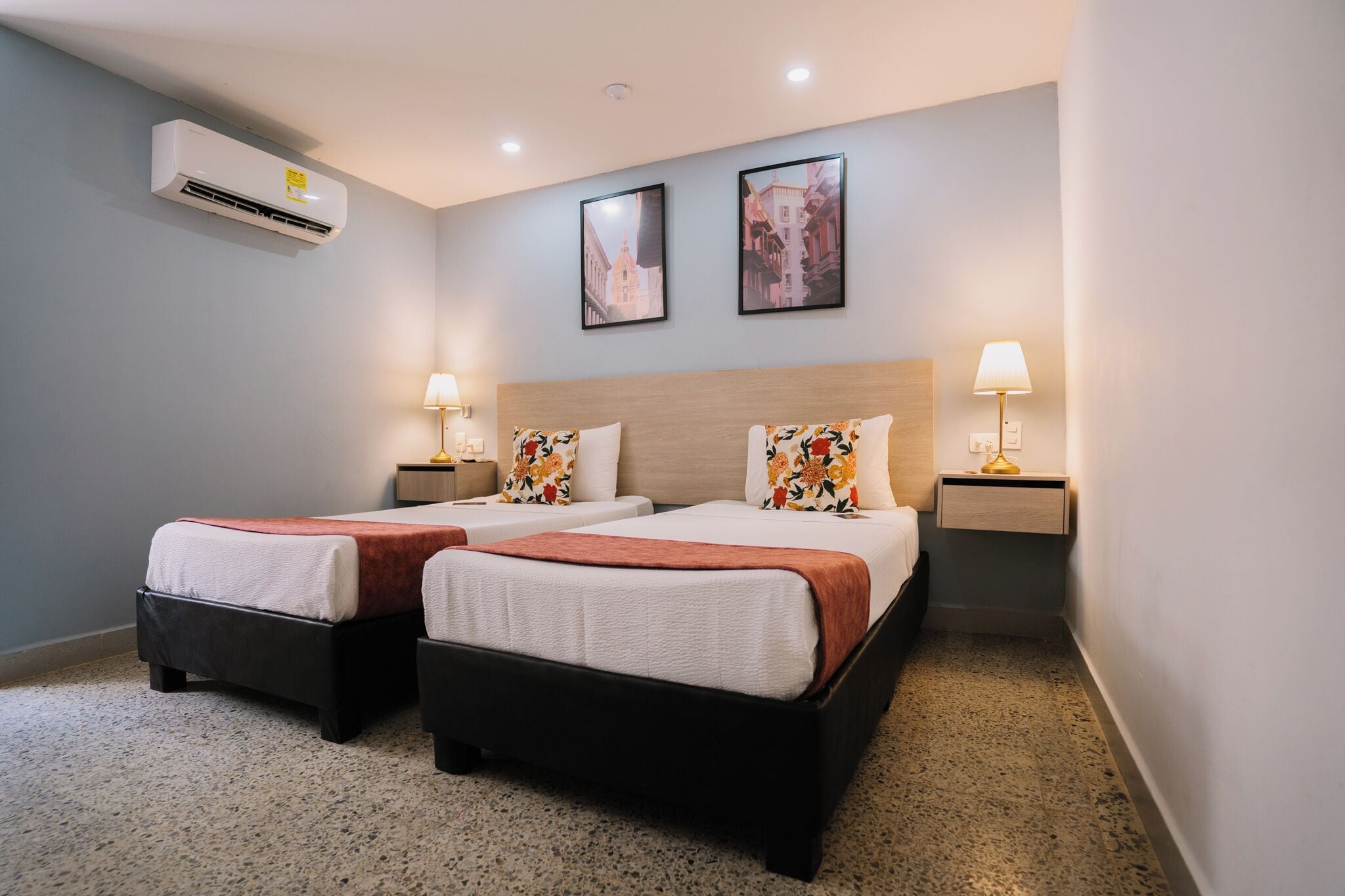 Ein Hotelzimmer mit zwei Betten, einer Wand-Klimaanlage, zwei gerahmten Bildern und Nachttischlampen.