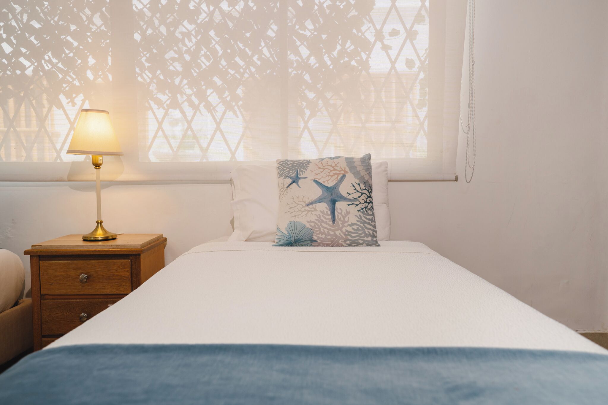 A neatly made bed with a white comforter and a blue throw blanket, a wooden nightstand with a lamp, and a decorative pillow with a sea-themed design.