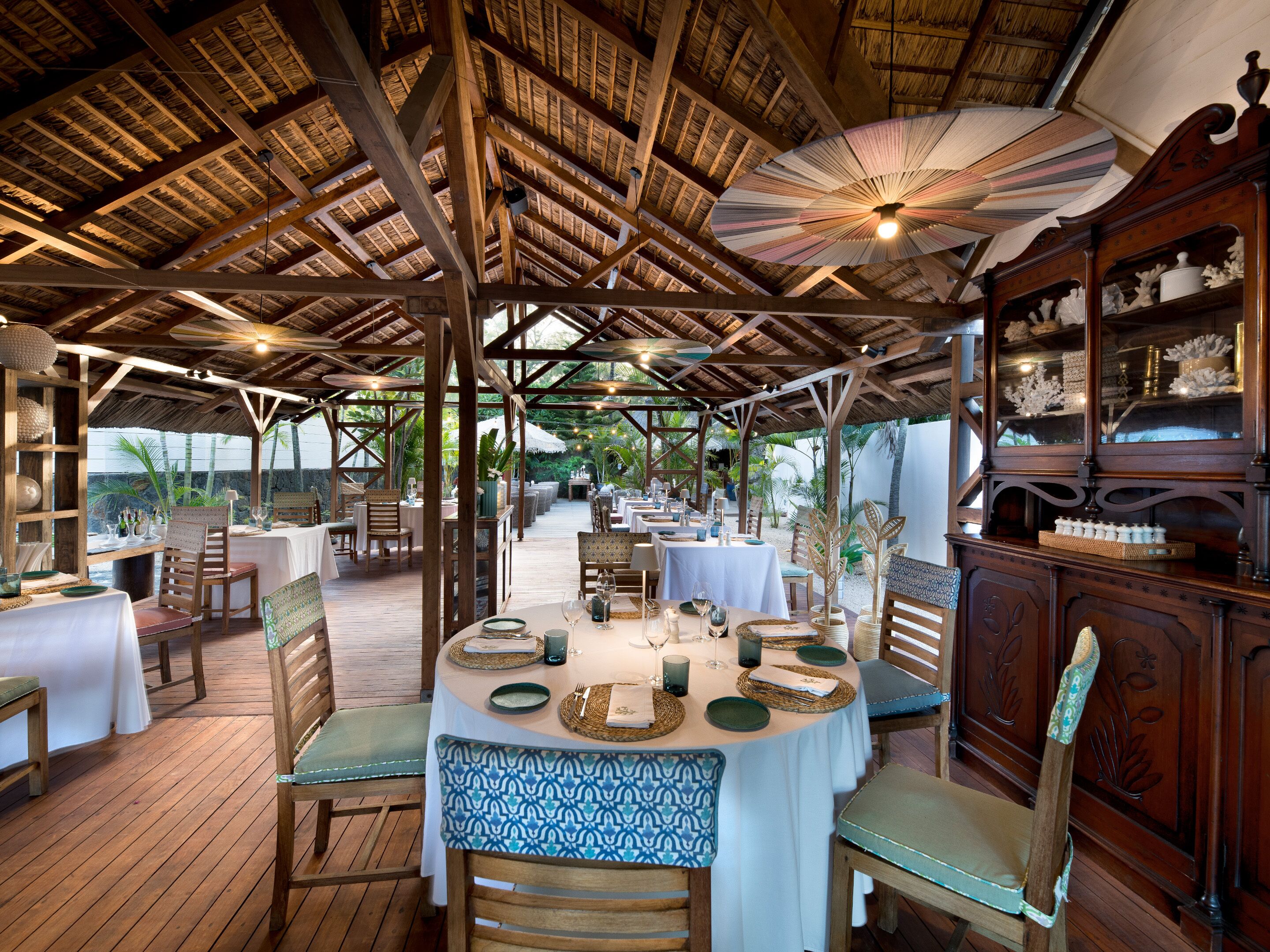 An outdoor dining area with wooden tables and chairs, a thatched roof, and a view of the surrounding landscape.