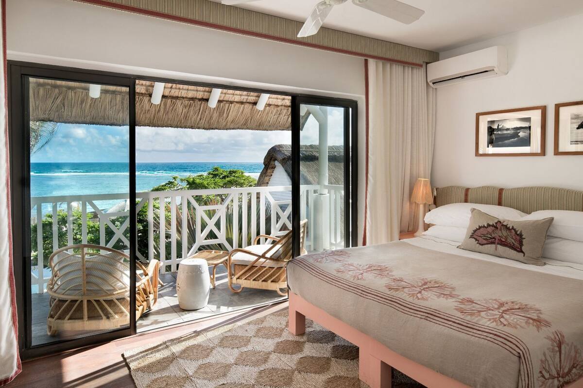 A bedroom with a bed, a ceiling fan, a nightstand, a lamp, and a view of the ocean through a sliding glass door.