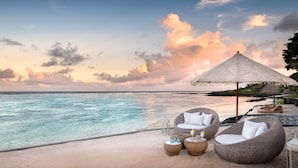 A beach setting with wicker lounge chairs, a thatched umbrella, and a view of the ocean.