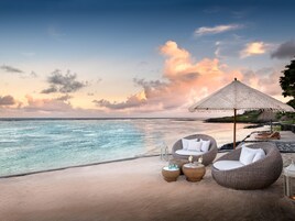 Escena de playa con sillones de mimbre, una sombrilla de paja y vista al océano.