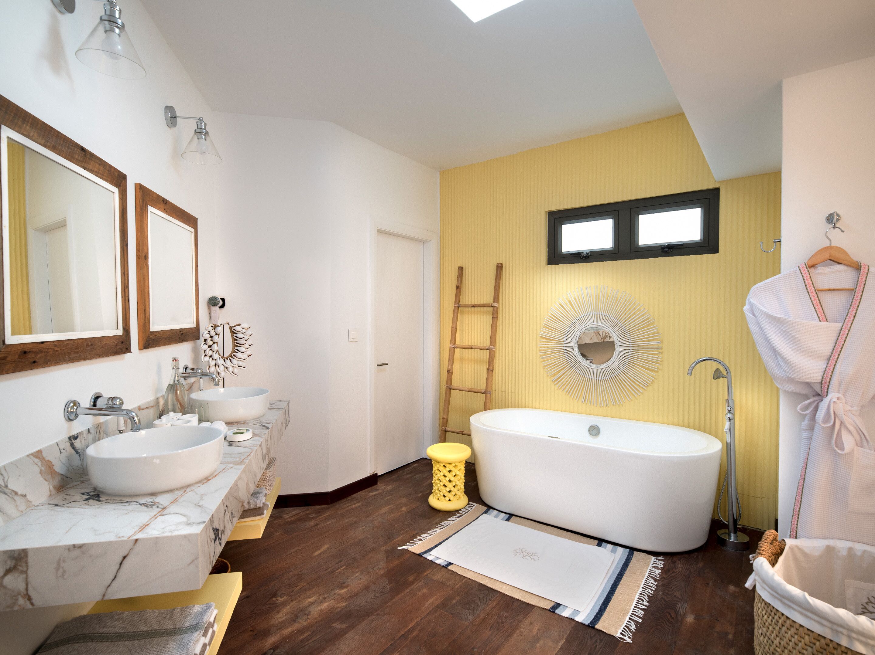A bathroom with a large bathtub, two sinks with marble countertops, a wooden ladder, and a sunburst mirror on the wall.