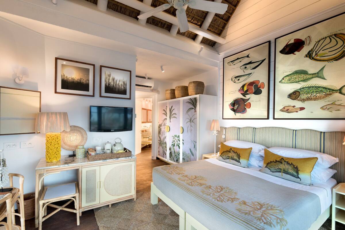 A bedroom with a bed, a desk, and a television. There are framed pictures on the wall and a ceiling fan.