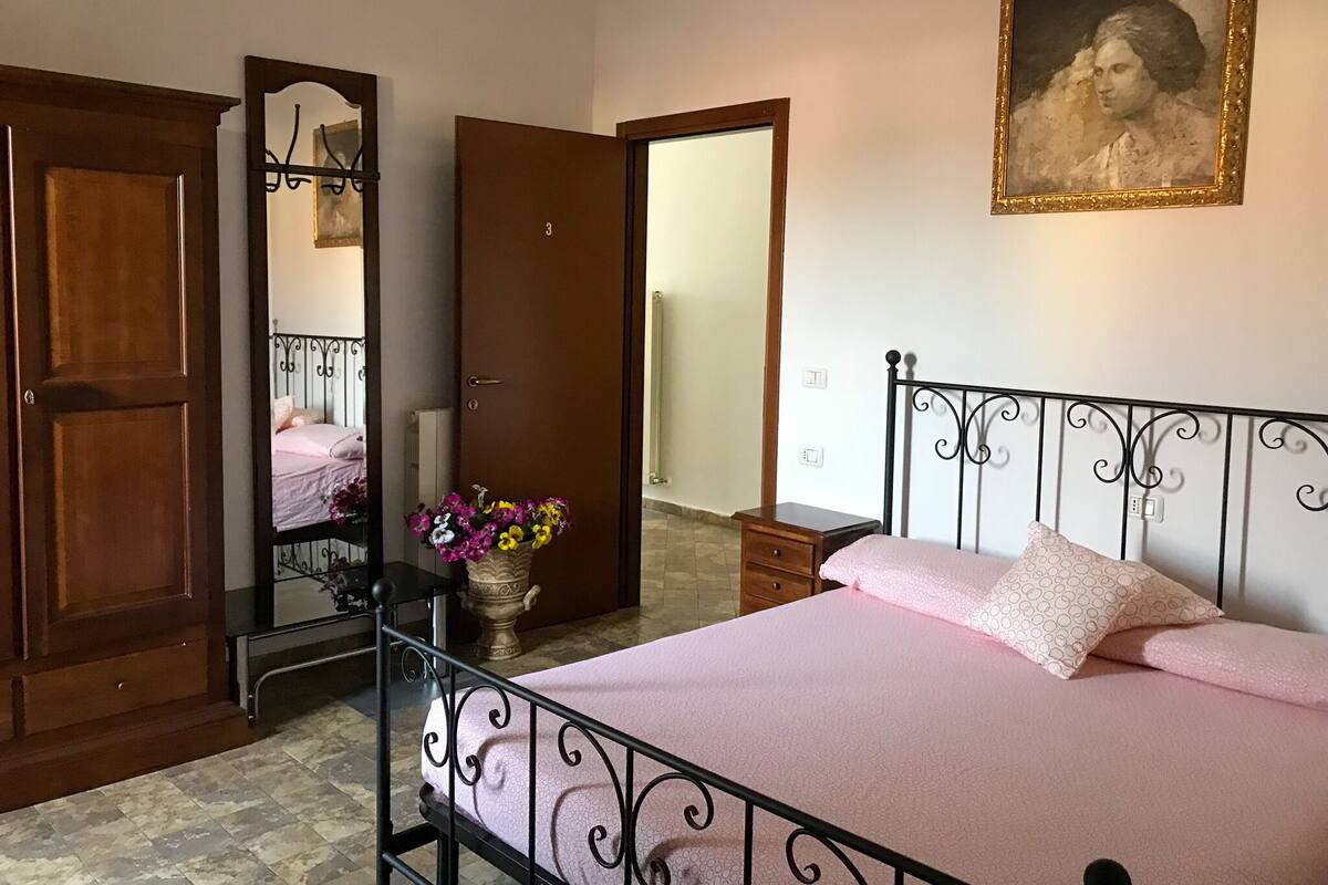 A bedroom with a bed, a wooden wardrobe, a mirror, a vase with flowers, and a painting on the wall.