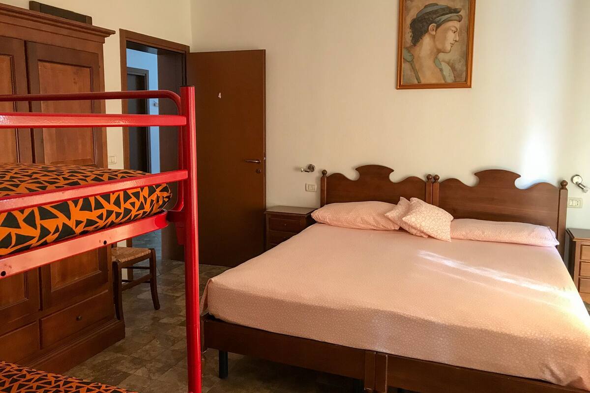 A bedroom with a bunk bed, a single bed, a wooden wardrobe, and a door leading to another room.
