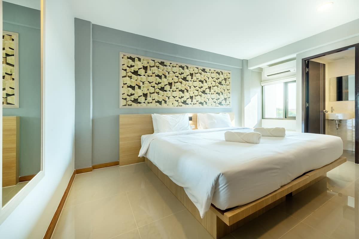 A modern hotel room with a large bed, a wooden headboard, a decorative wall panel, and a bathroom with a large mirror and sink.