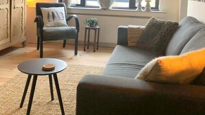 A cozy living room with a grey sofa, a small round table, a chair, and a cabinet. There is a large window with a plant on the windowsill.