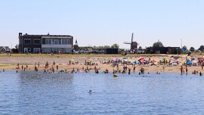 A beach with people swimming and sunbathing, a windmill, and buildings in the background.