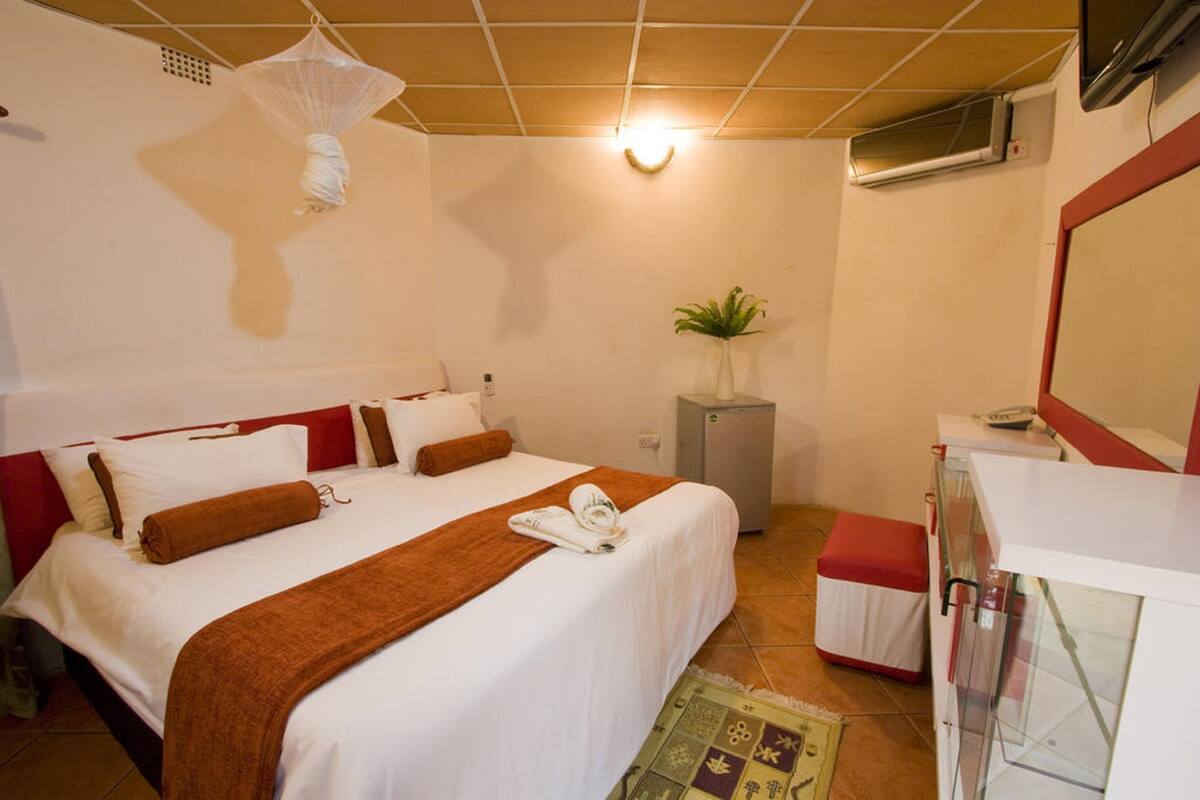 Quarto de hotel com uma cama, uma pequena escrivaninha, uma planta e um espelho.