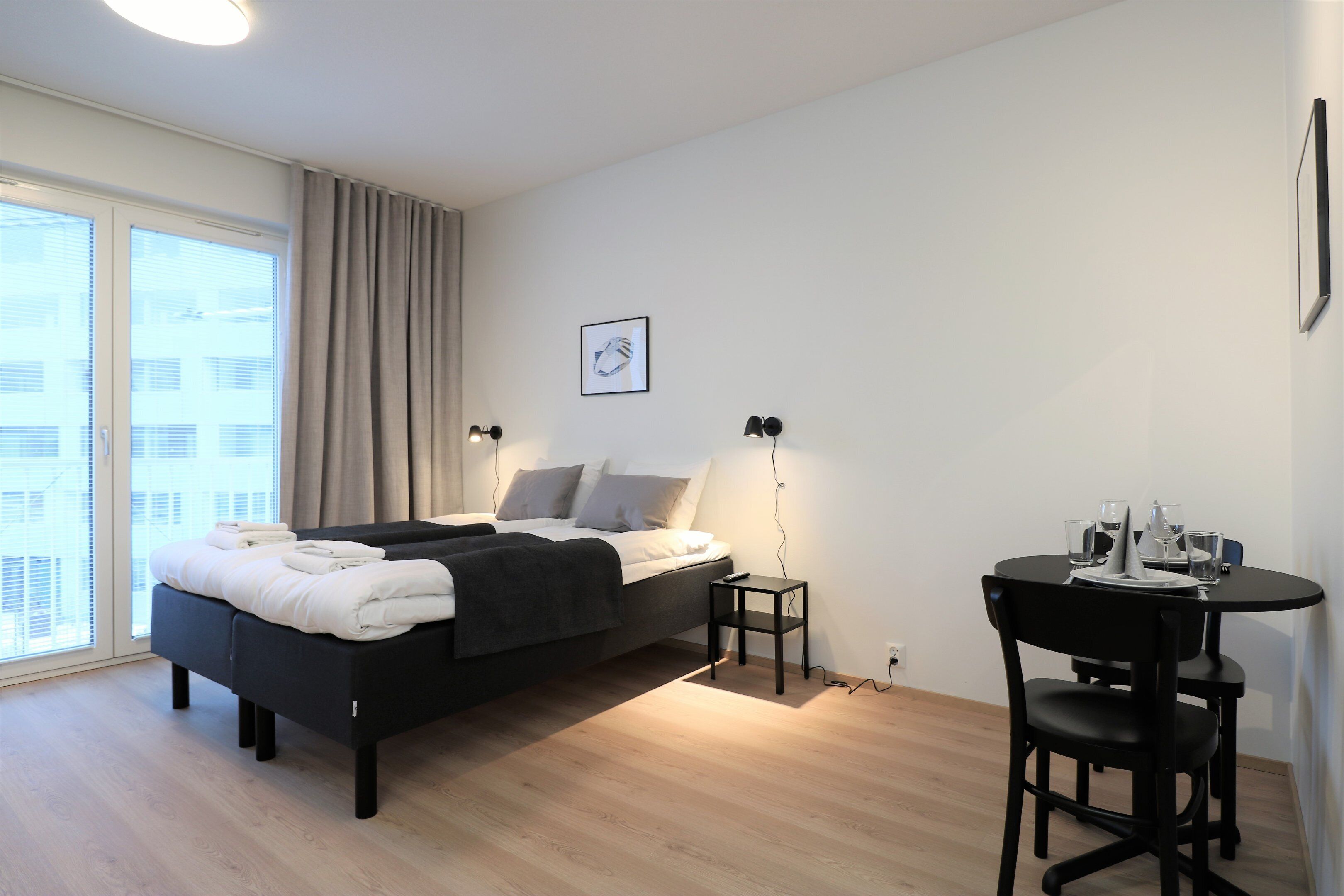 Une chambre à coucher moderne, avec un lit, des tables de chevet, une petite table avec des chaises et une fenêtre avec des rideaux.