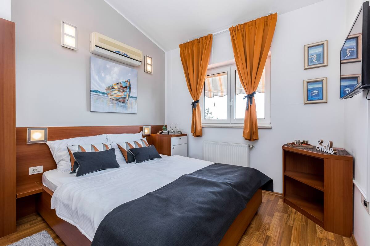 A hotel room with a bed, a nightstand, a dresser, a window with curtains, a wall-mounted TV, and artwork.