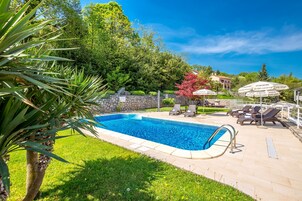 Una piscina con sillas de descanso y sombrillas, rodeada de follaje y un muro de piedra.