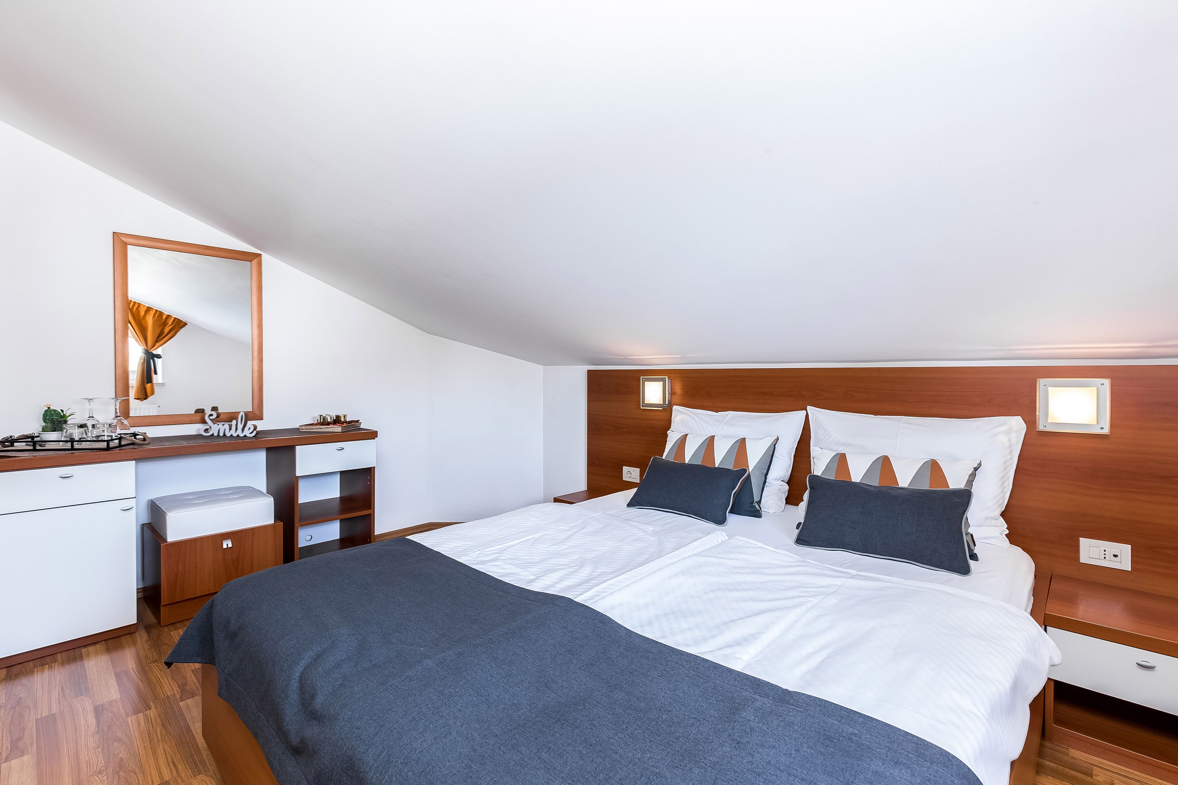 Een moderne slaapkamer met een houten hoofdbord, een groot bed met wit beddengoed, een nachtkastje en een dressoir met spiegel.