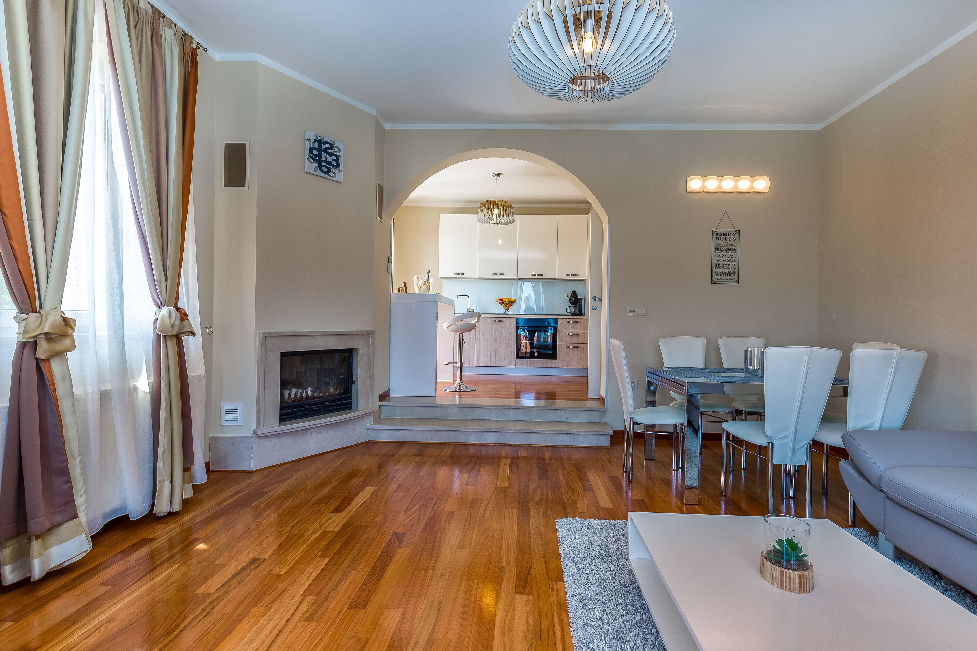 Una sala moderna con chimenea, piso de madera, una zona de comedor con sillas blancas y una cocina al fondo.