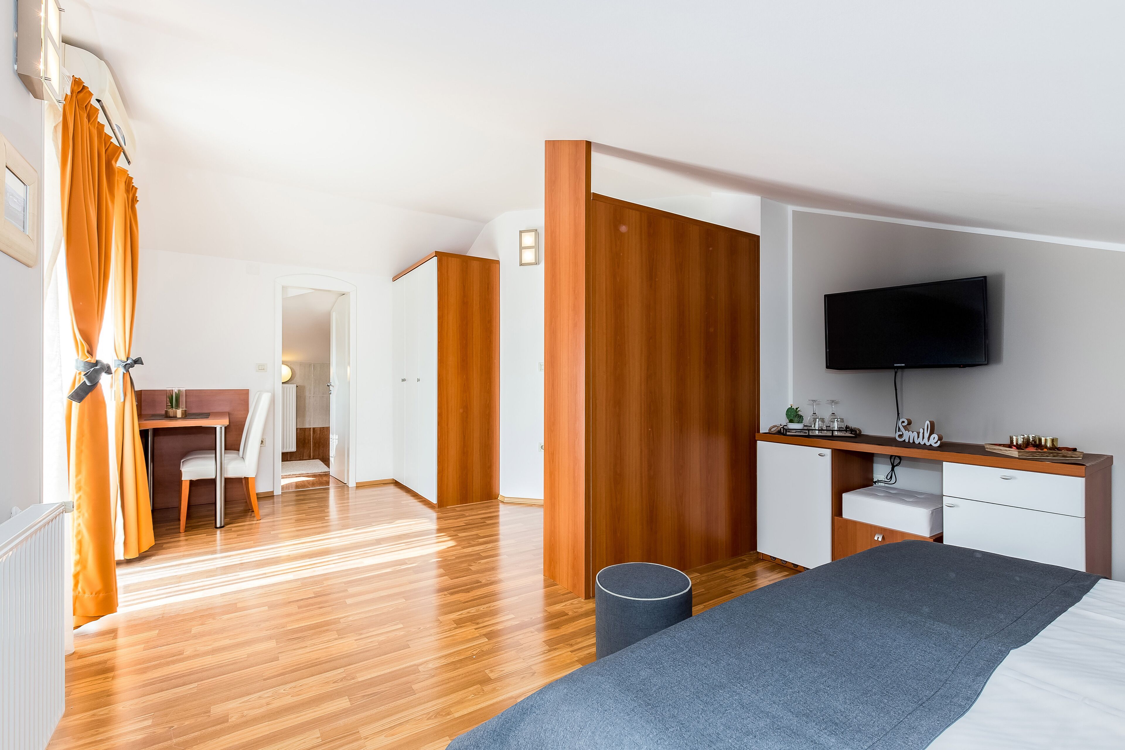 Une chambre d’hôtel moderne avec un plancher en bois, un lit recouvert d’une couverture grise, un bureau avec une chaise, une armoire et un téléviseur fixé au mur.