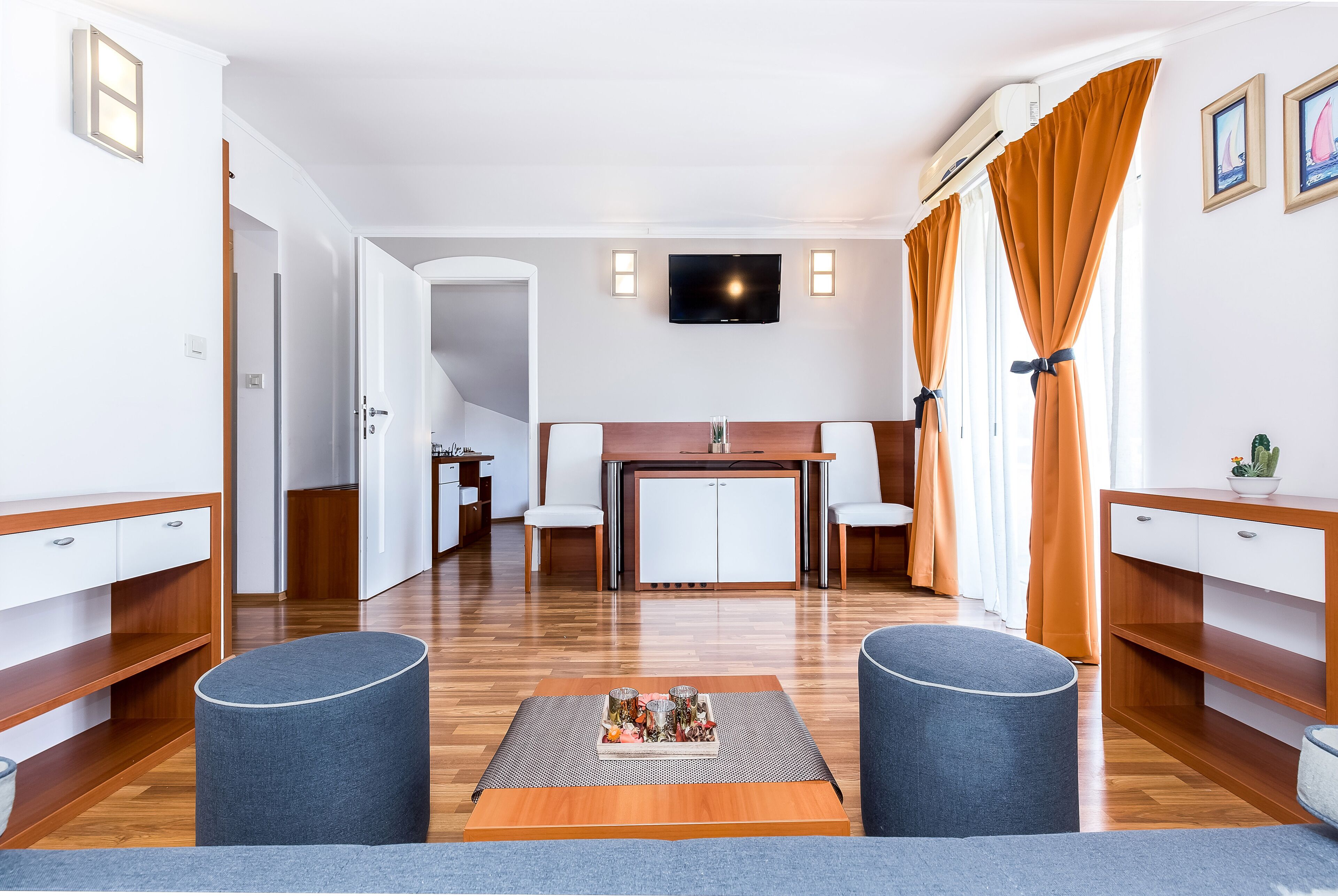 Una habitación de hotel moderna con suelo de madera, una zona de comedor con mesa y sillas, un televisor montado en la pared y dos otomanas.