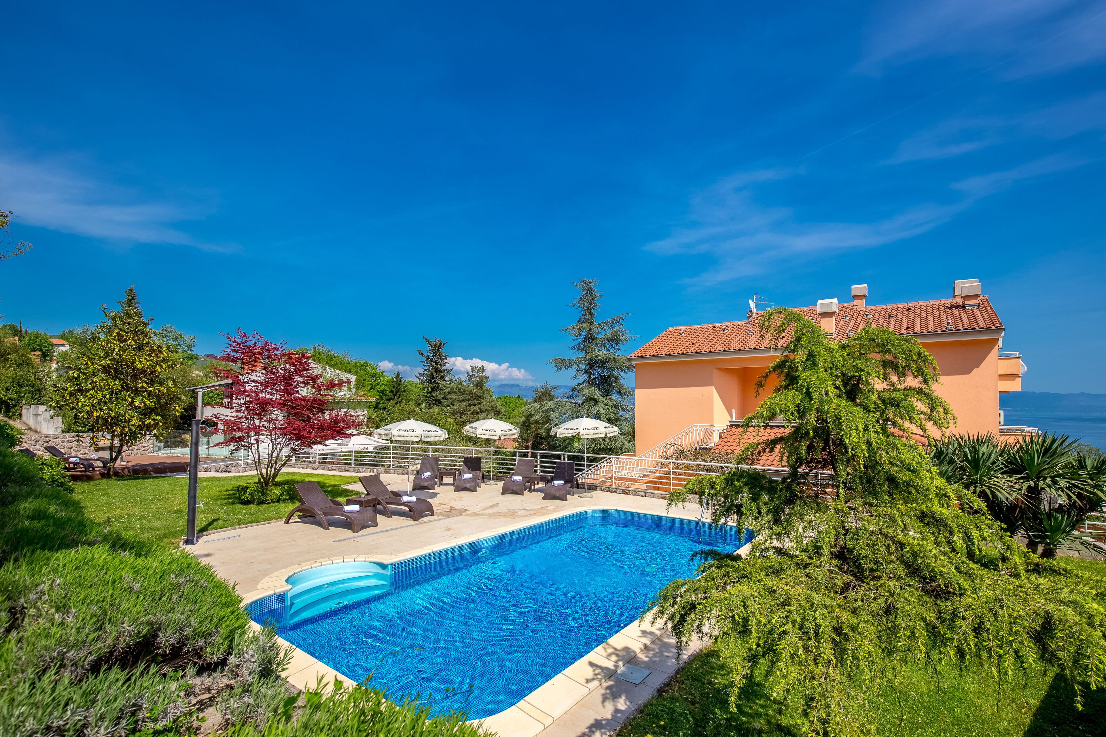 A swimming pool with lounge chairs and umbrellas, surrounded by greenery and a building with a red-tiled roof.