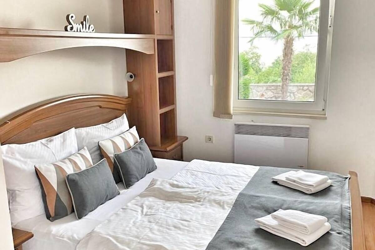 A neatly made bed with a wooden headboard, a shelf with a 'Smile' sign, and a window with a view of palm trees.