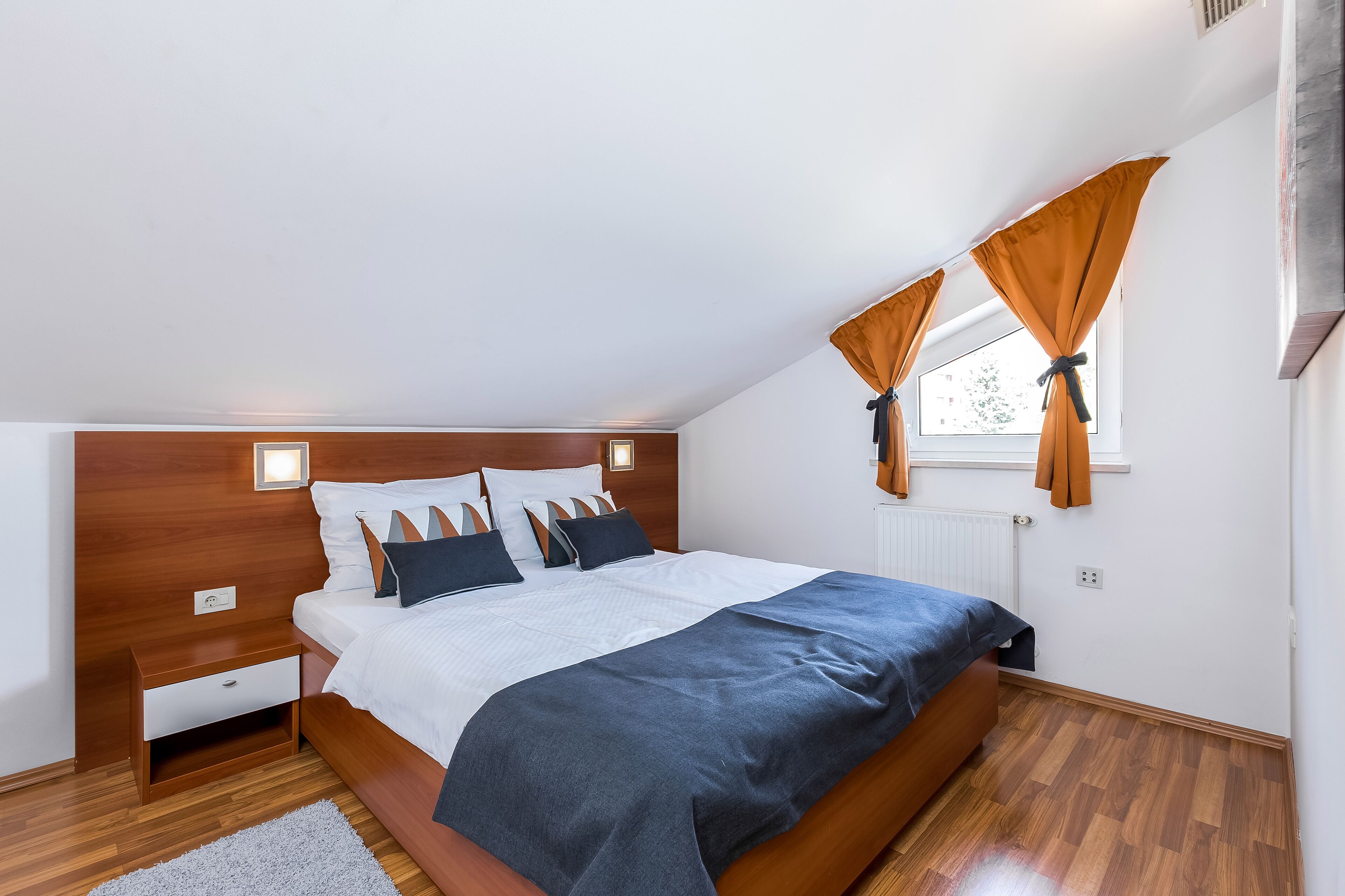 A modern bedroom with a wooden headboard, a large bed with a dark blue bedspread, white bedding, and two bedside tables.
