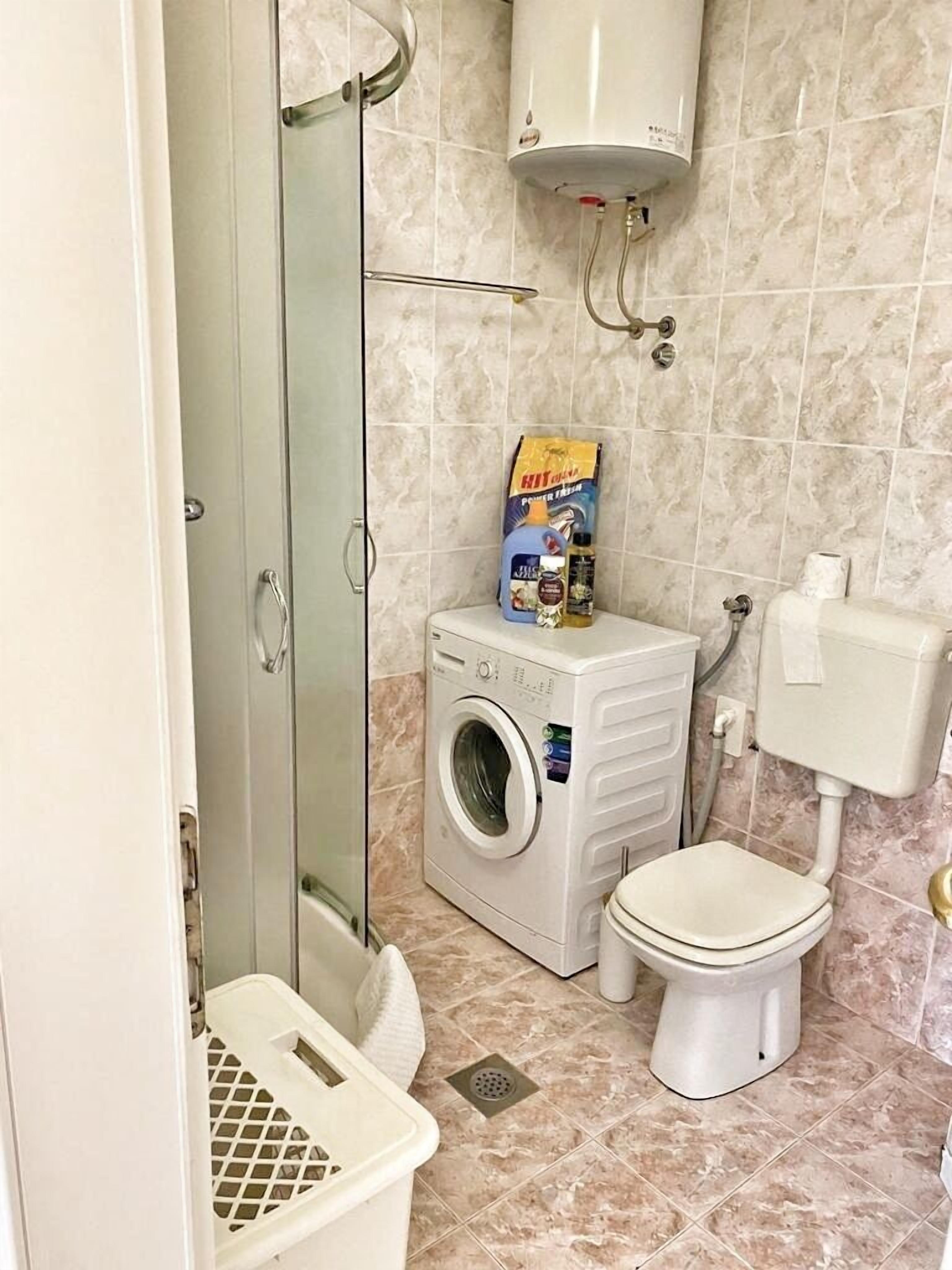 A compact bathroom with a glass shower enclosure, a washing machine, a toilet, and a small laundry basket.