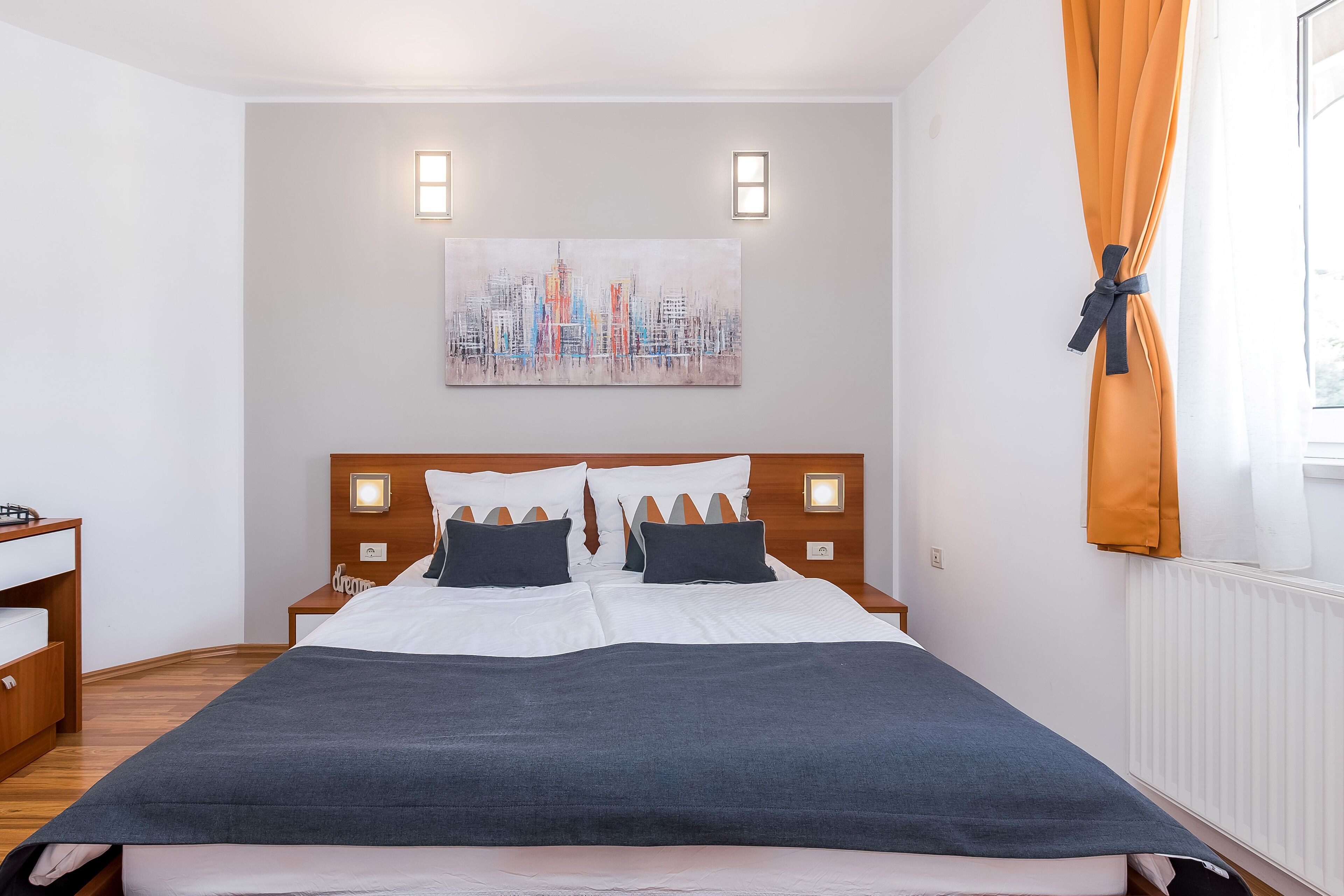 A hotel room with a neatly made bed, a cityscape painting on the wall, and a window with curtains.