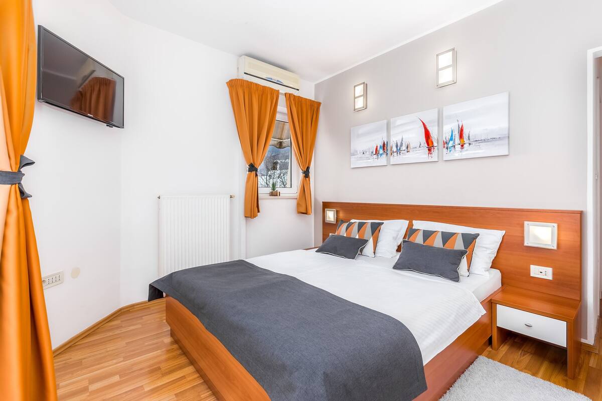 A modern bedroom with a wooden headboard, a flat-screen TV mounted on the wall, and a window with orange curtains.