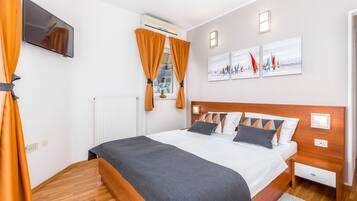 A modern bedroom with a wooden headboard, a flat-screen TV mounted on the wall, and a window with orange curtains.
