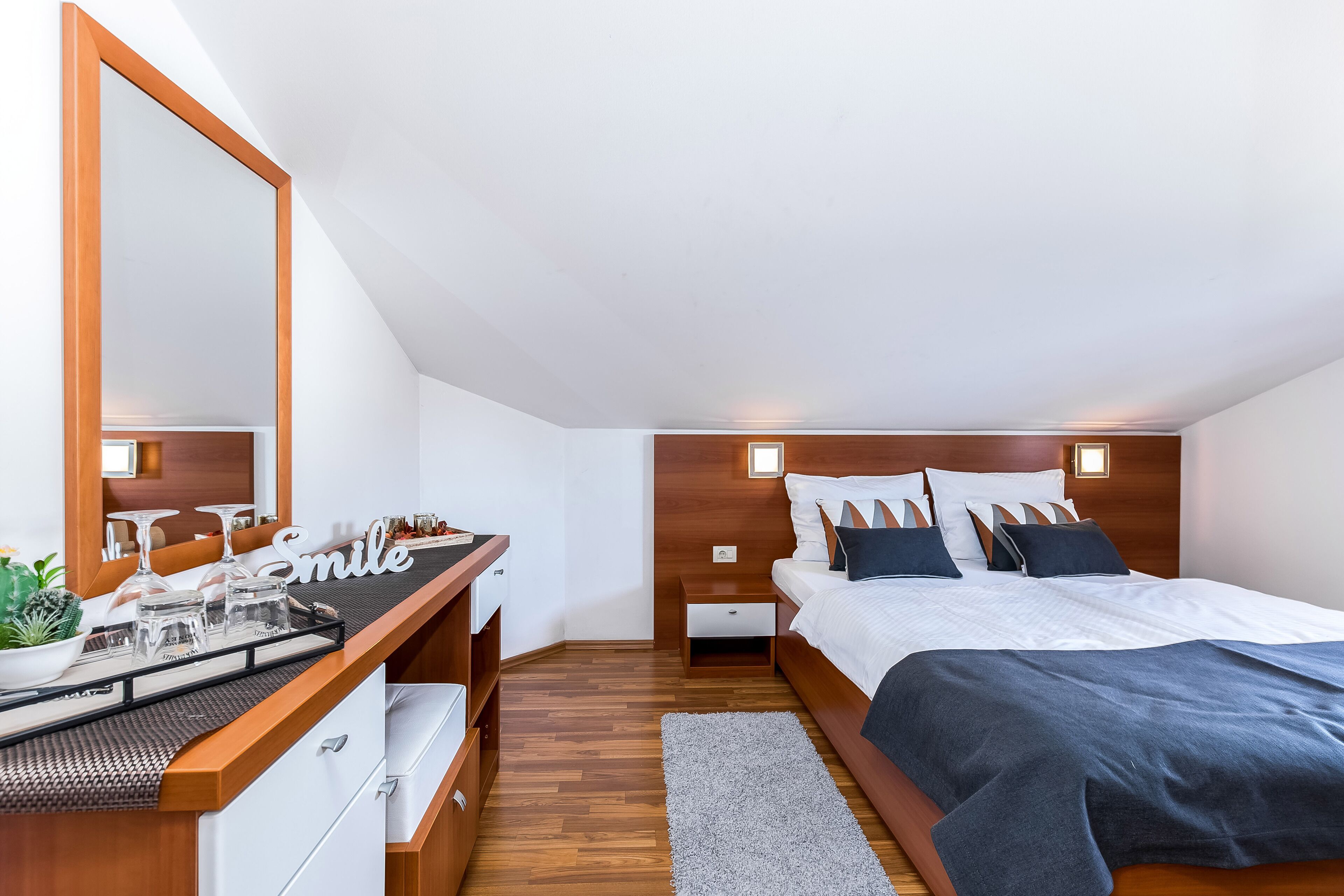 Un dormitorio con un escritorio de madera, un espejo grande, una cama con ropa oscura y una mesita de noche pequeña.