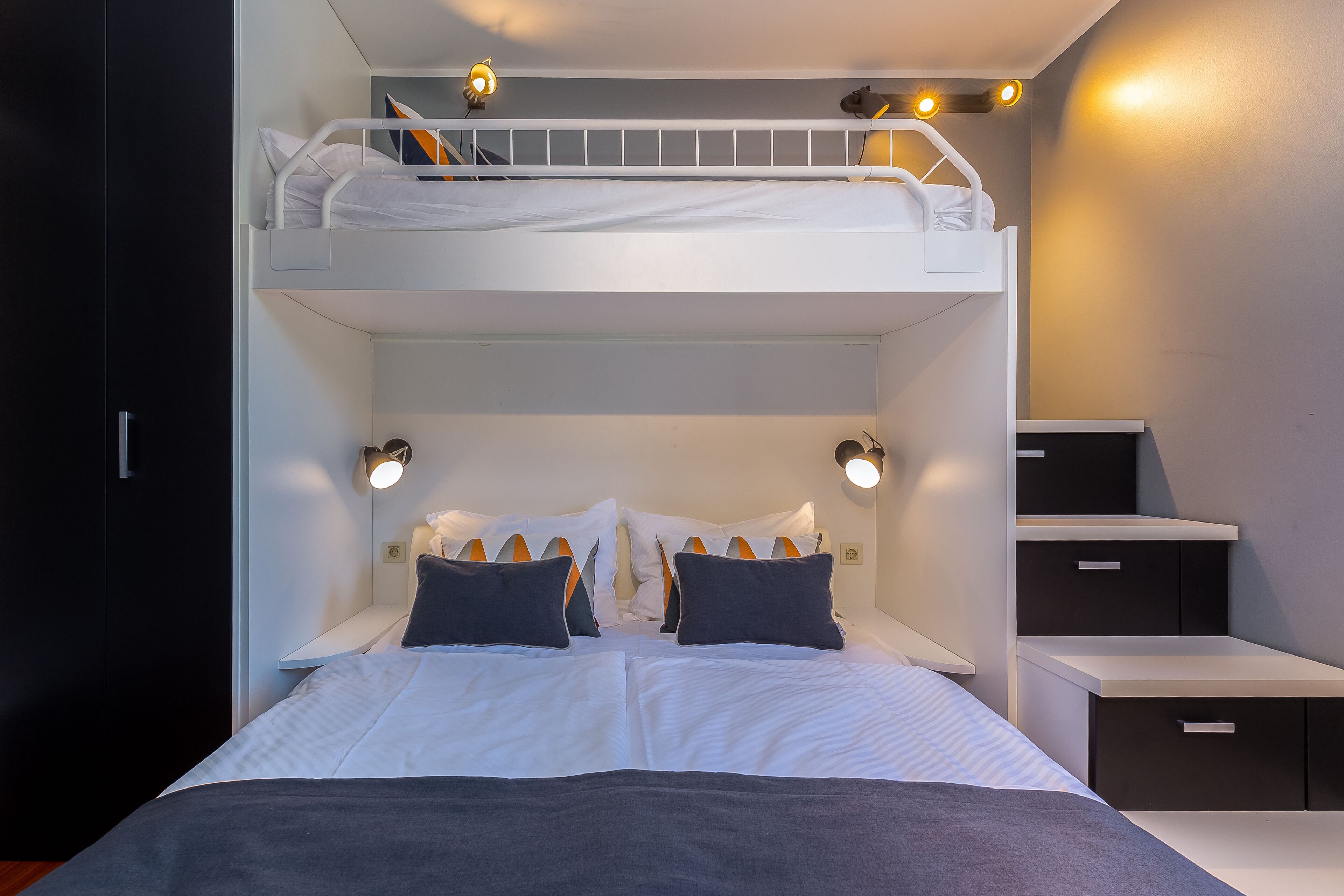 A bunk bed room with a desk and nightstands, white bedding, and wall-mounted reading lights.