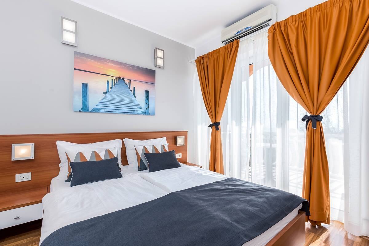 A modern hotel room with a wooden headboard, a large bed with white and dark blue bedding, a sunset painting on the wall, and large windows with orange curtains.