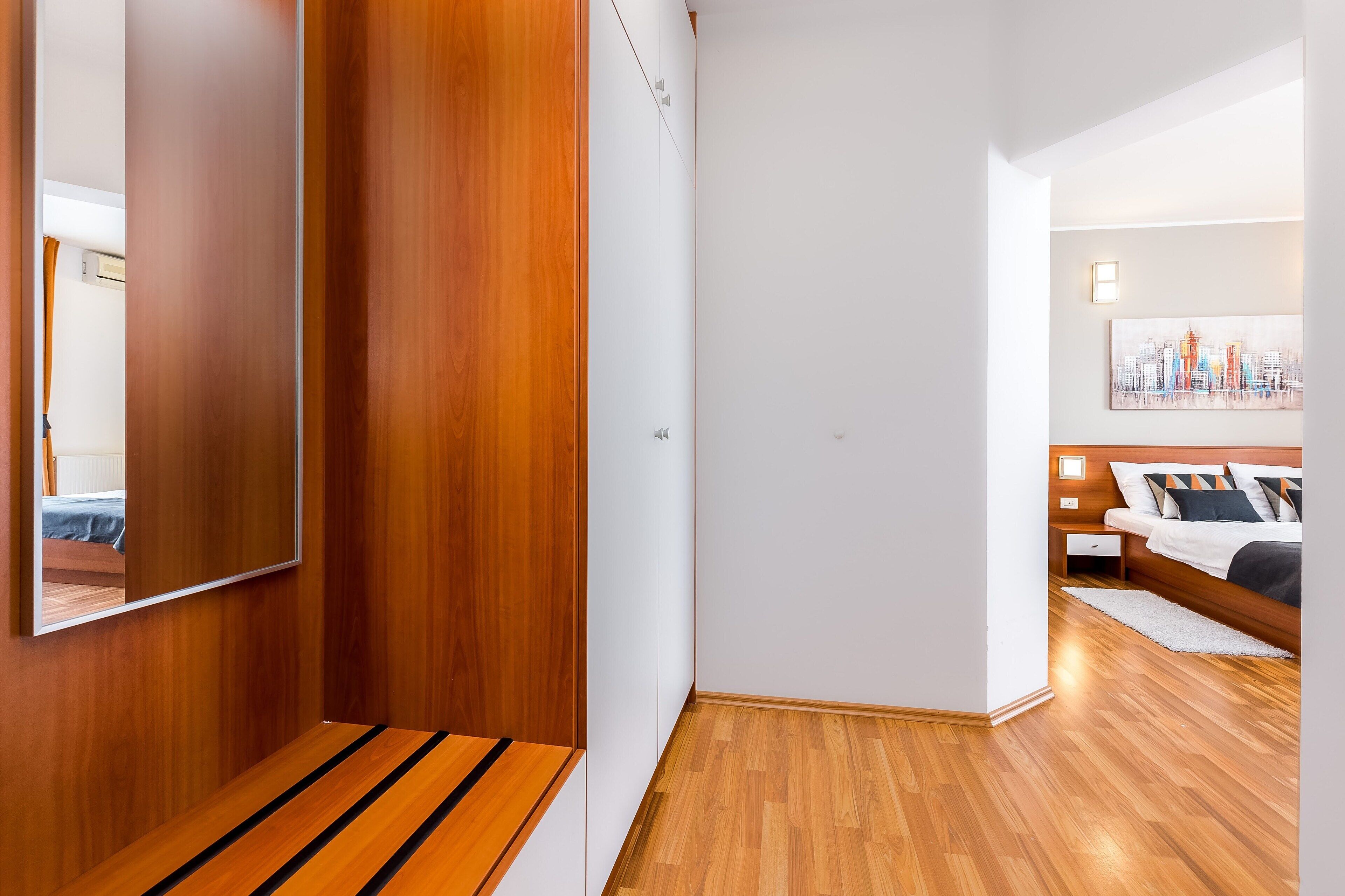 A modern hotel room with a wooden floor, a bed with a white headboard, a nightstand, and a wall-mounted mirror.
