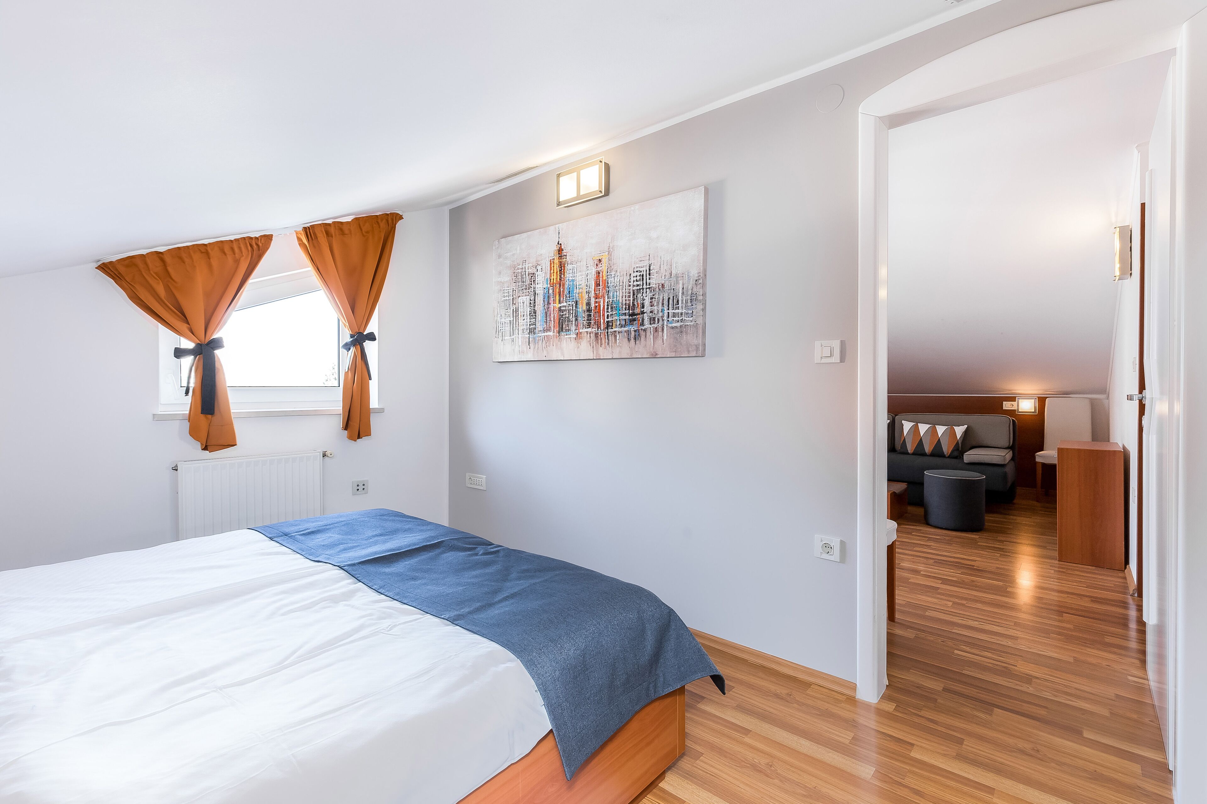 A bedroom with a bed, curtains, a cityscape painting, and a hallway leading to a living area.