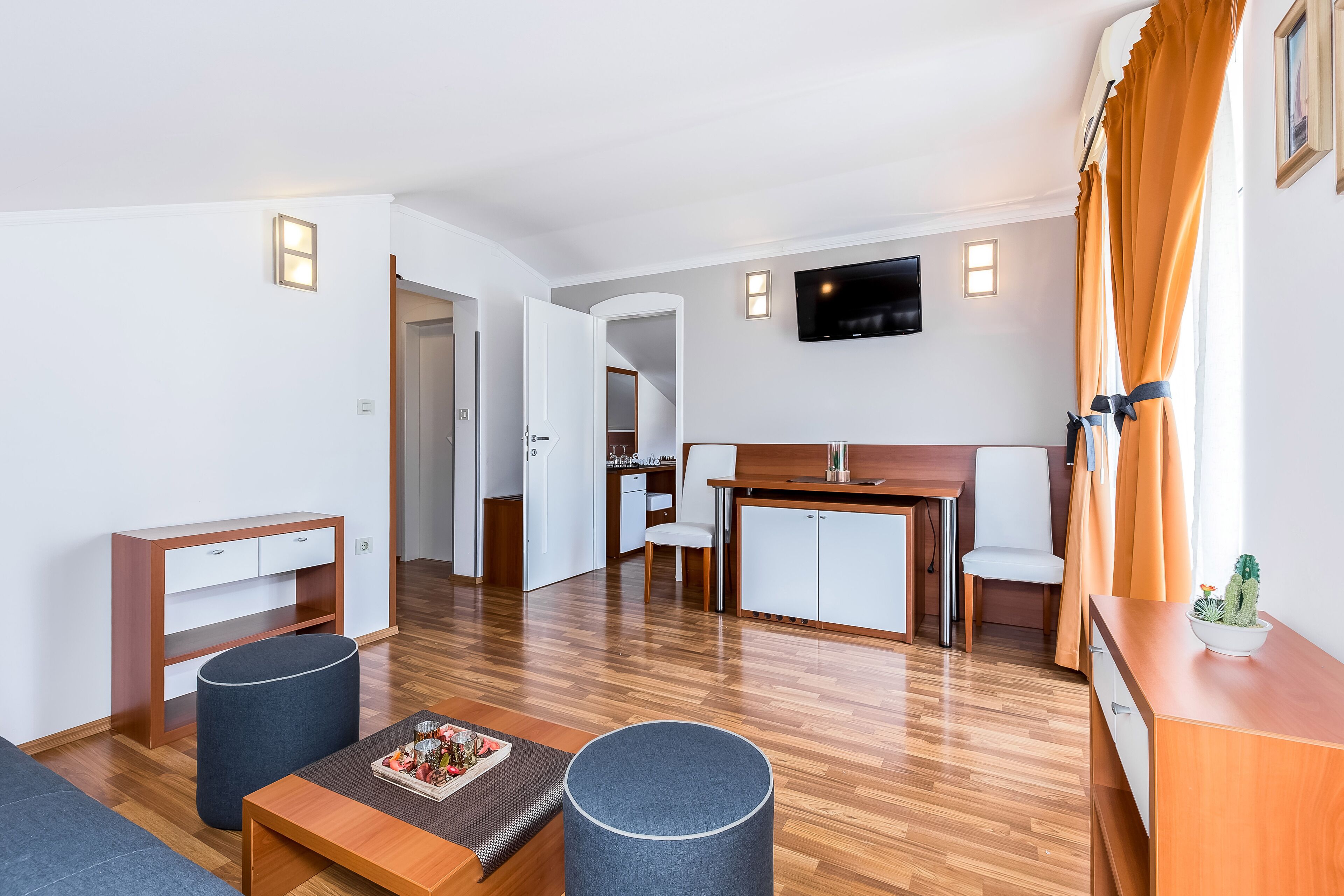 A modern hotel room with a wooden floor, a sofa, a small table, a TV, and a kitchen area with a dining table and chairs.