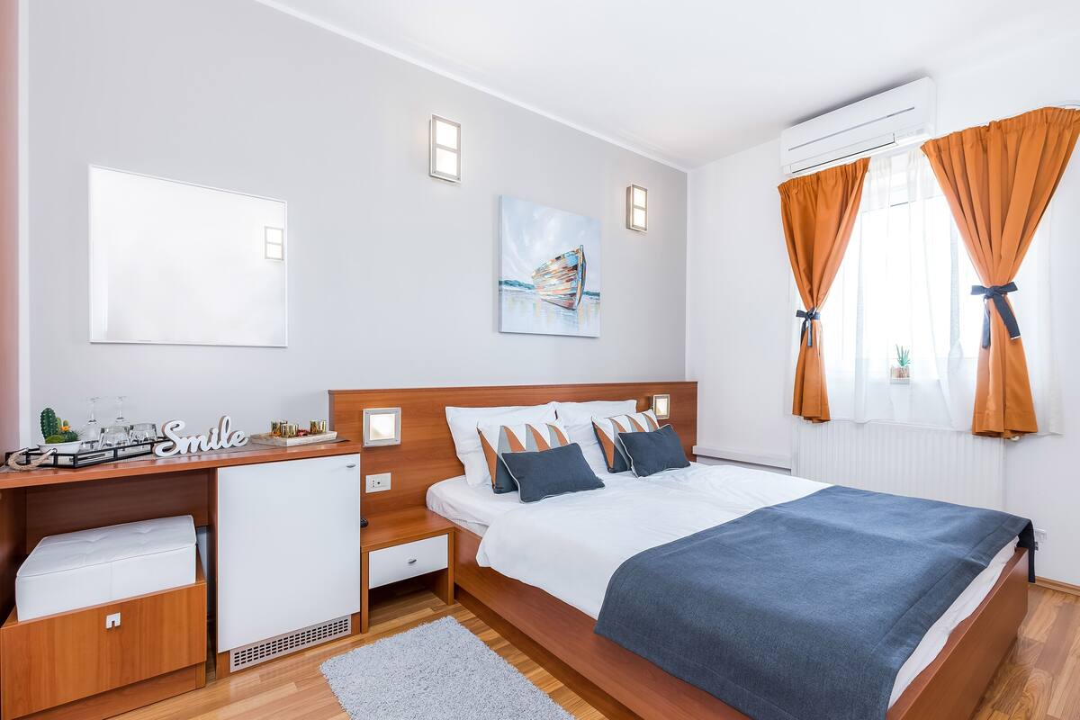 A modern bedroom with a wooden bed, white walls, a desk with a computer, and a small refrigerator.