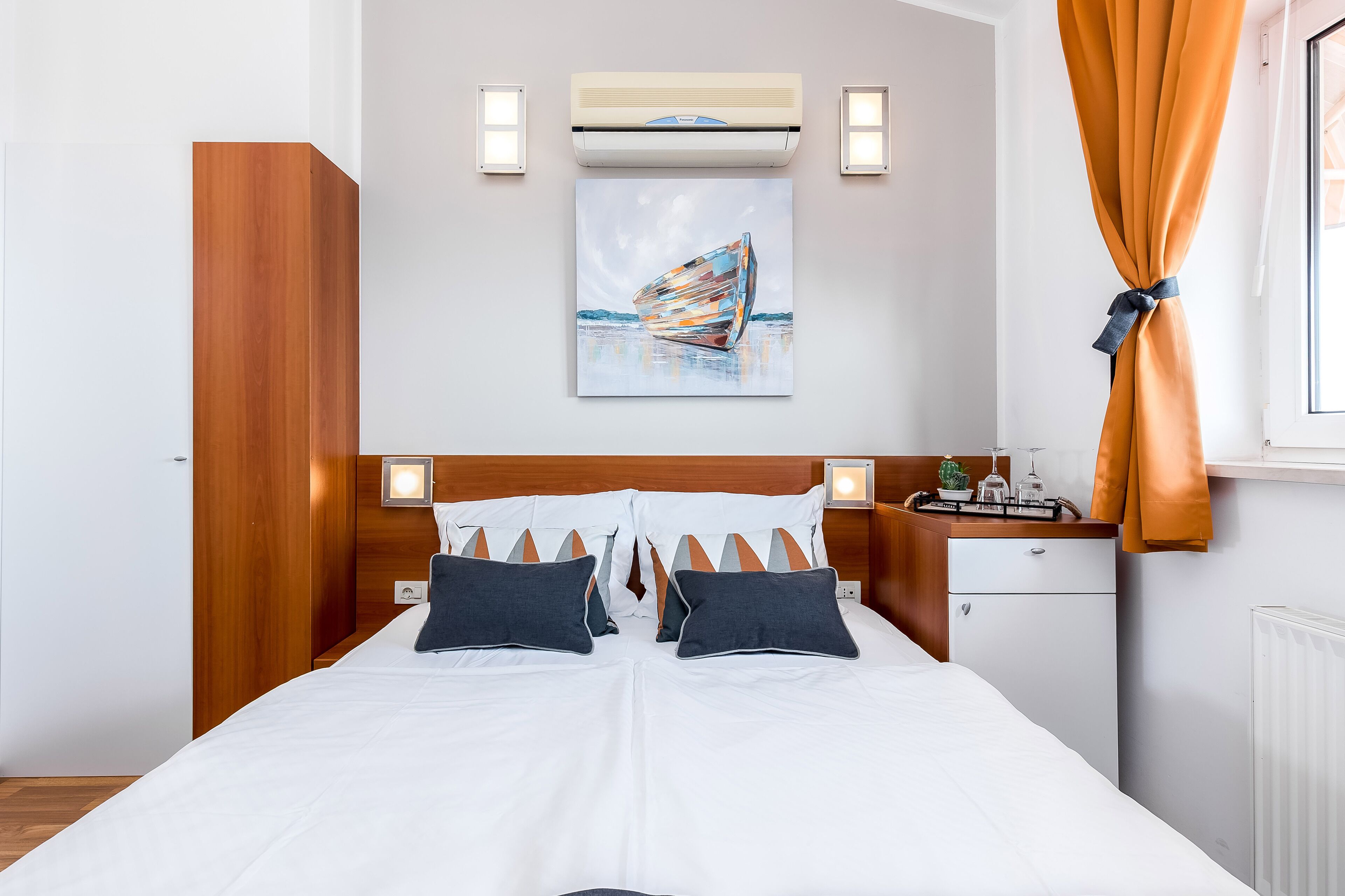 A neatly made bed with white linens and two dark pillows, a wooden headboard, a small bedside table with a lamp, and a painting of a boat on the wall.