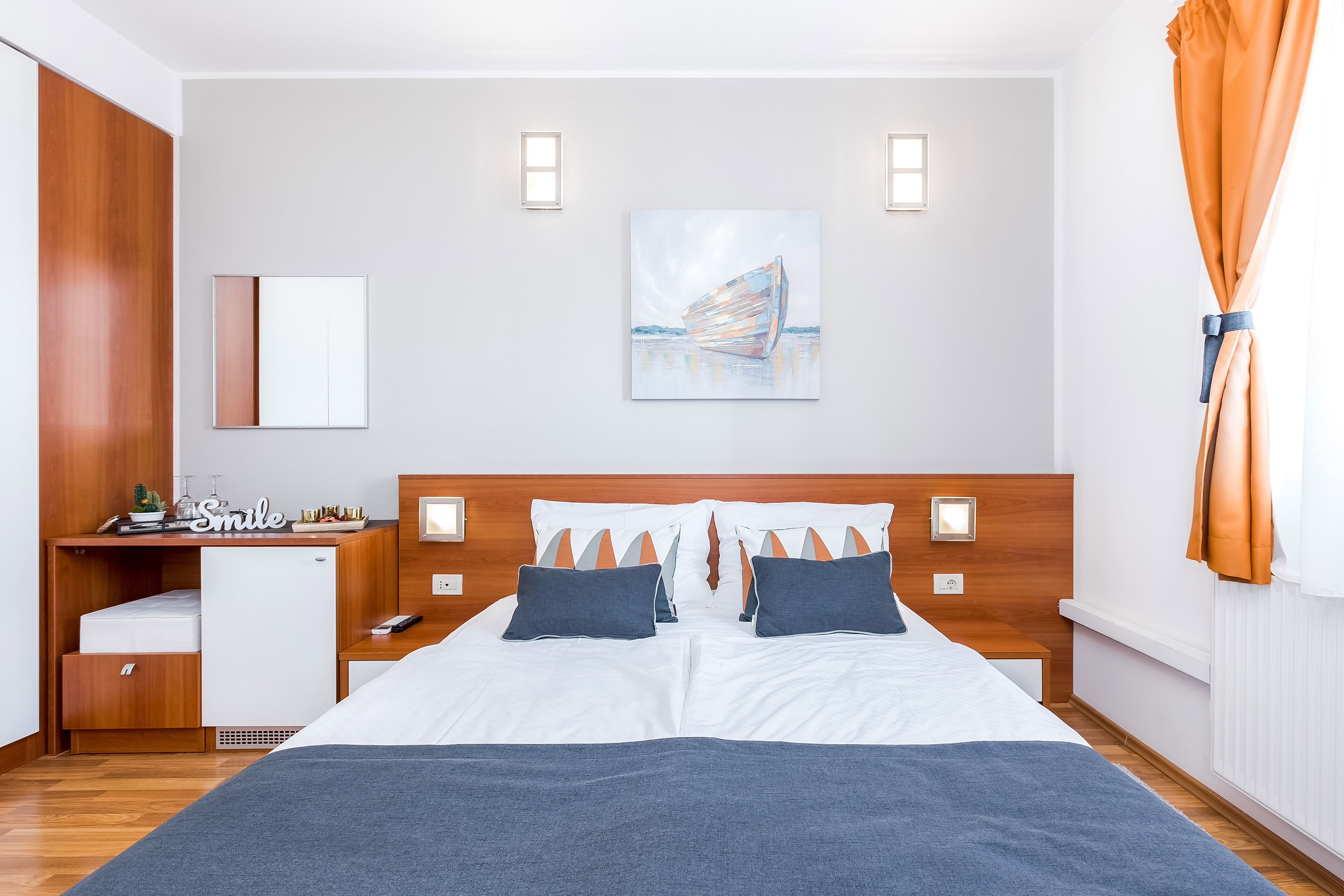 A hotel room with a bed, bedside tables, a desk with a chair, a small refrigerator, and a window with curtains.