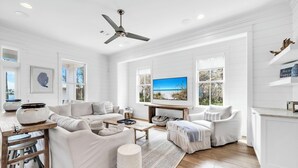 A spacious living room with a large TV, white furniture, and a ceiling fan.