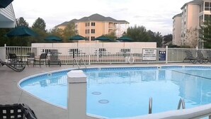 A swimming pool with lounge chairs and umbrellas, surrounded by a white fence and buildings in the background. - At home feeling with amenities. (Branson)