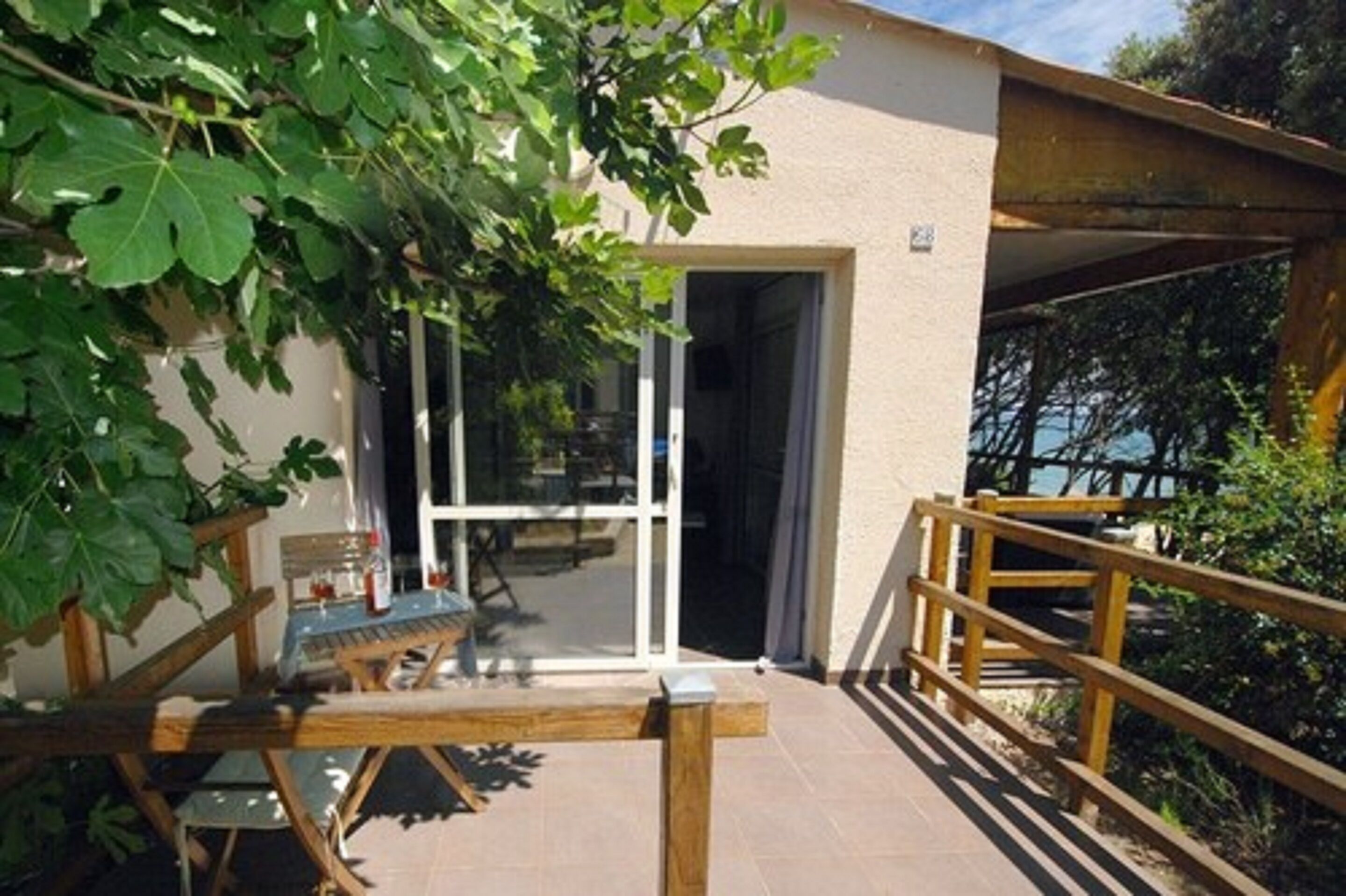 Een terras met een houten tafel en stoelen, een glazen deur die naar een binnenruimte leidt, en een houten balustrade.