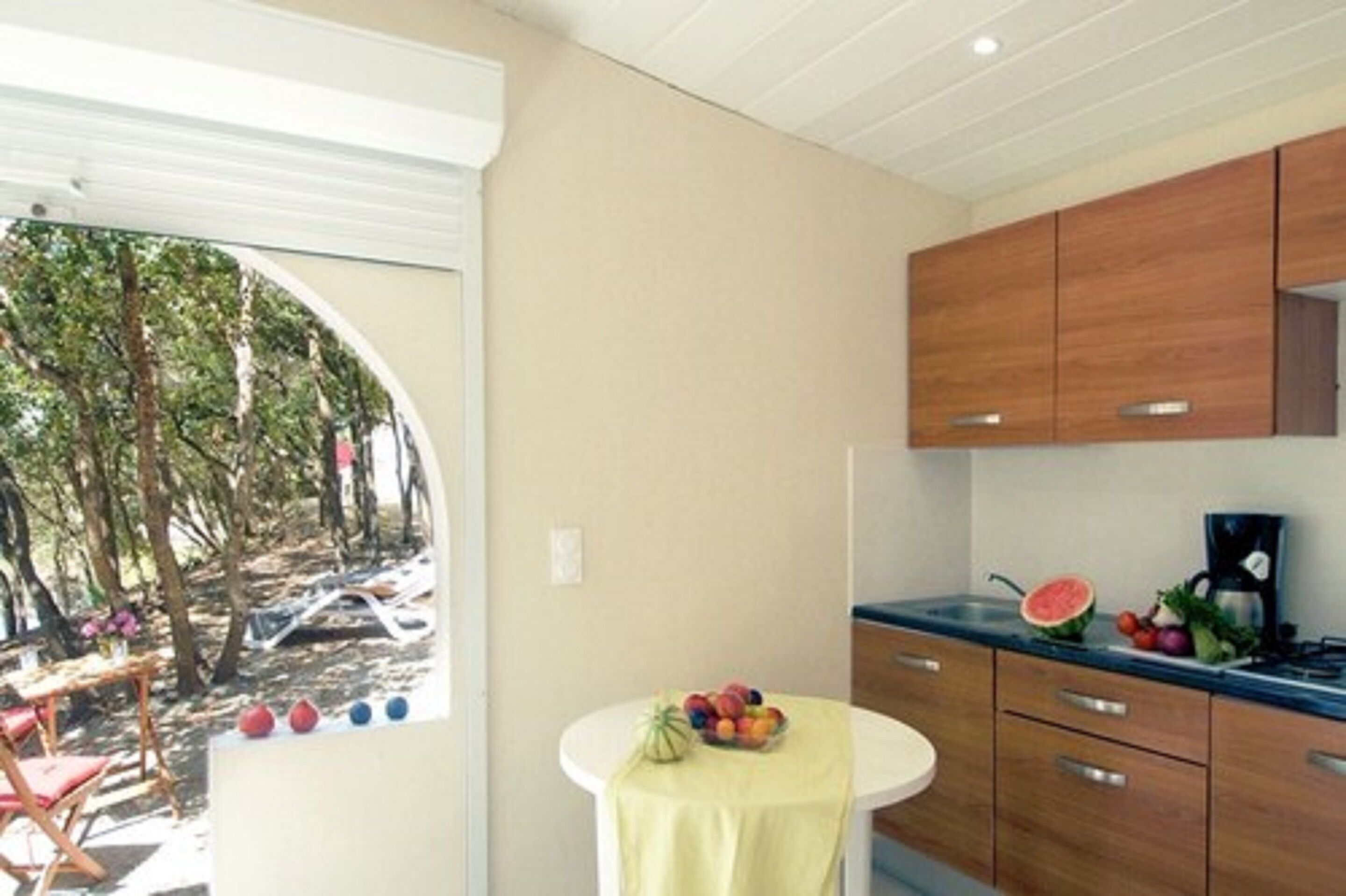 Een moderne keuken met houten kastjes, een ronde tafel met een schaal fruit en uitzicht op bomen.