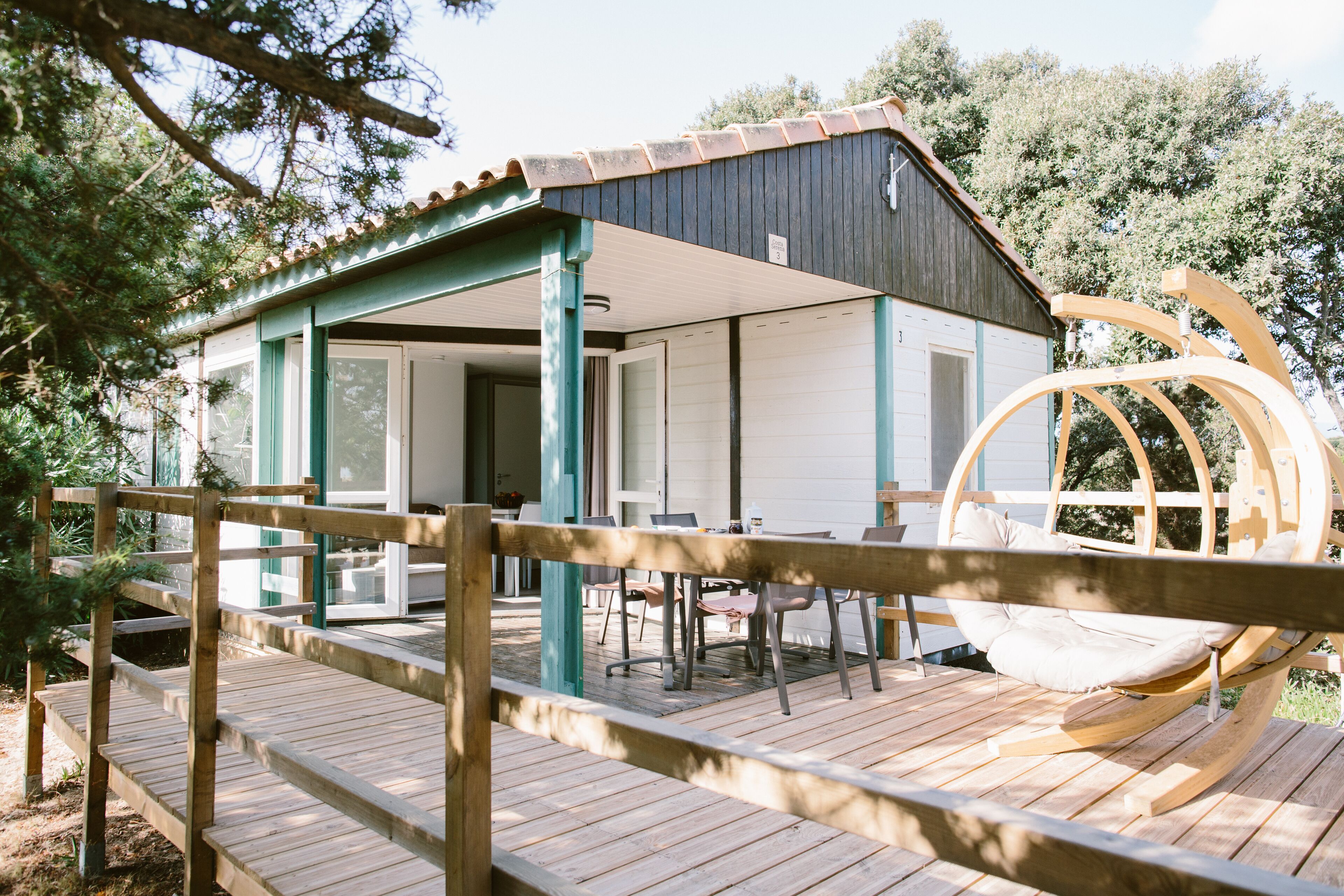 Une terrasse en bois avec un fauteuil suspendu, un espace extérieur aménagé et couvert, et un bâtiment doté d’un toit sombre et de murs blancs.