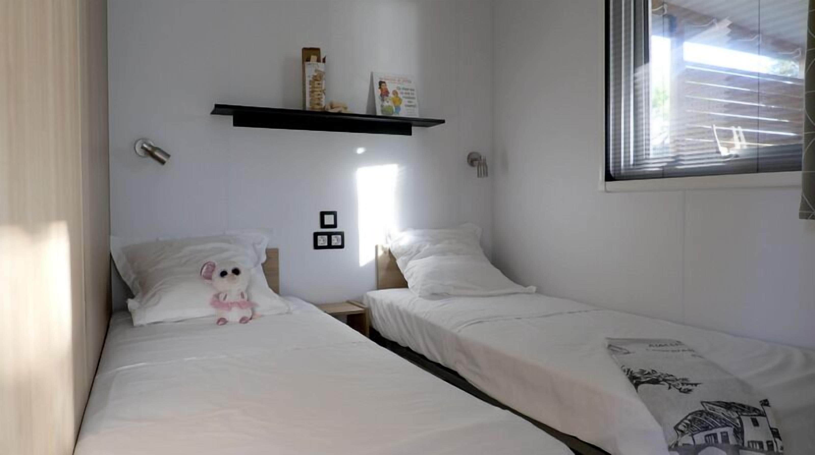 A bedroom with two single beds, a shelf with books and a framed picture, and a window with blinds.