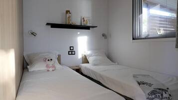 A bedroom with two single beds, a shelf with books and a framed picture, and a window with blinds.