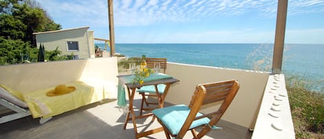 A patio with a table and chairs overlooking the sea, a small building, and a covered area with a hat and towel.