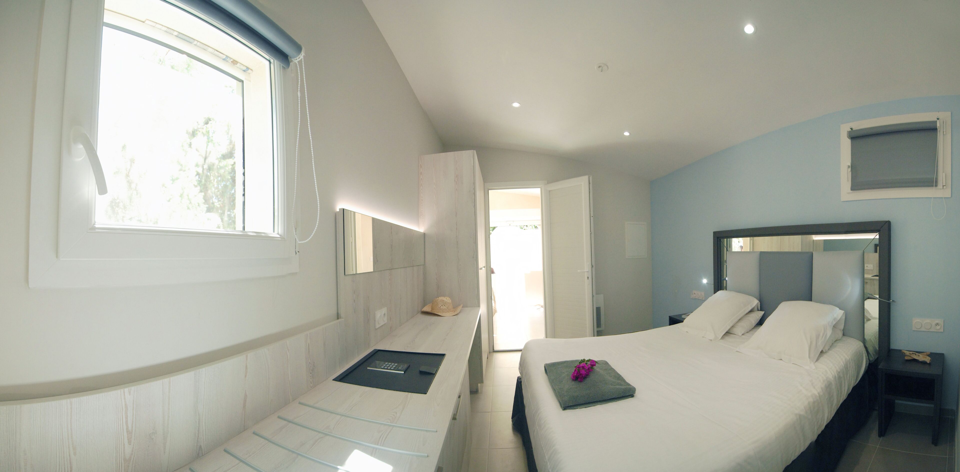 A bedroom with a bed, a bedside table, a window, and a door leading to another room.